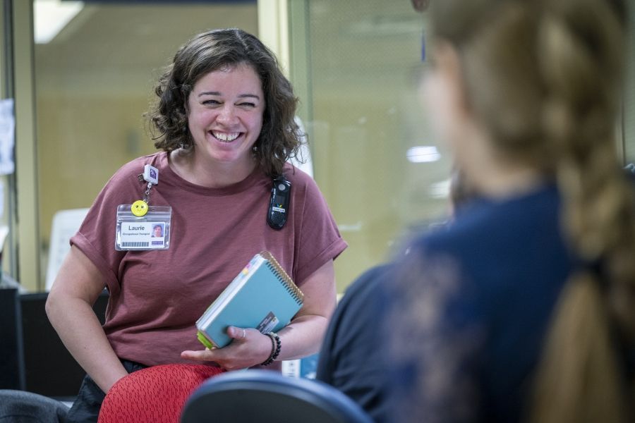 PeopleofKHSC_Laurie_Hill_6 Laurie Hill is smiling and laughing while chatting with a colleague, whose back is facing the camera. Hill is holding her notebooks. She has short brown wavy hair and is wearing a dusty rose blouse and black pants.