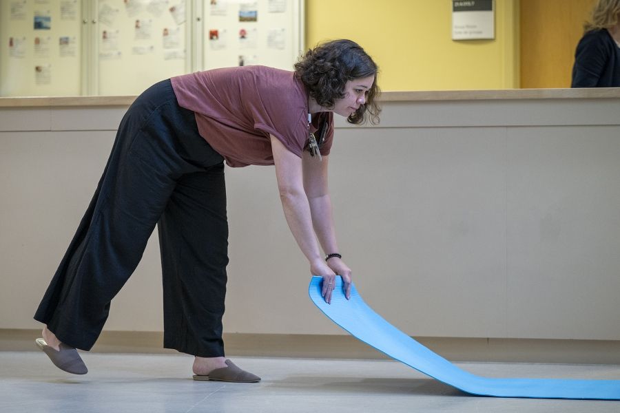 PeopleofKHSC_Laurie_Hill_7 Laurie Hill is unrolling a blue yoga mat and placing it on the floor. She has short brown wavy hair and is wearing a dusty rose blouse and black pants.