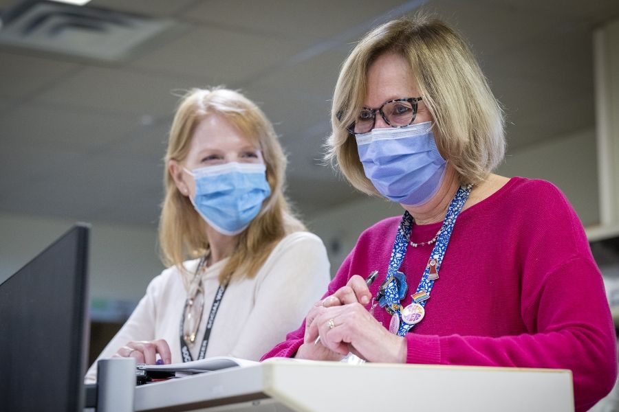 PeopleOfKHSC_Clare_Bowley_5 A wide shot of Clare Bowley and another social worker not in focus, standing and chatting at a care desk with masks one. Clare has a blonde bob, blue eyes, has tortoise framed glasses on, and is wearing a pink sweater with her decorated KHSC lanyard on top.