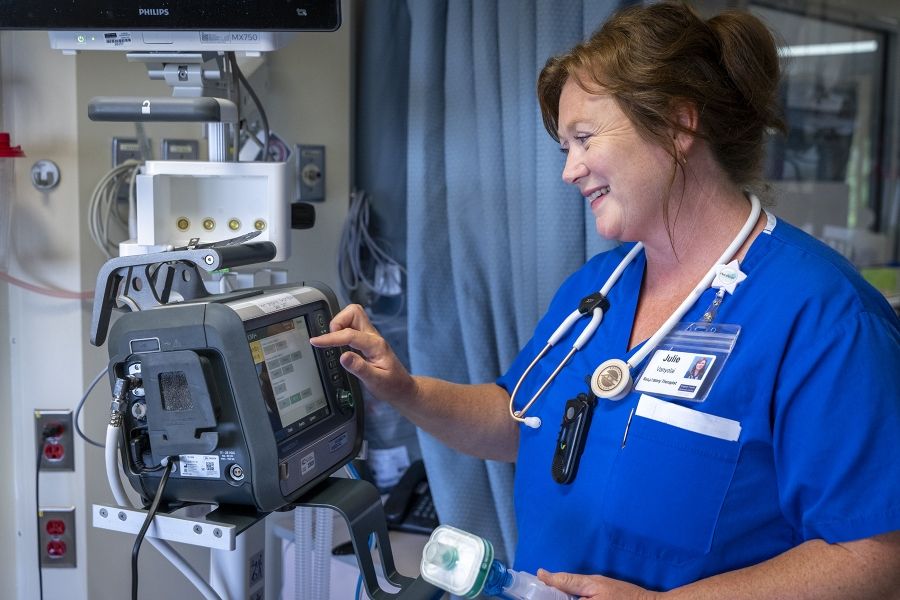 Julie Vanyolai is pictured standing, facing a piece of equipment. She’s tapping a button and holding a suction tube. She has strawberry blonde/red hair, which is tied up in a bun, and blue eyes. She’s wearing blue scrubs and has a stethoscope around her neck.