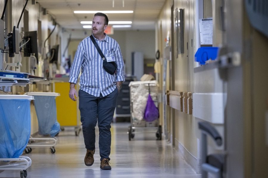 PeopleOfKHSC_Khayman_Wood_4 Khayman Wood is walking down a hallway at the Kingston General Hospital site. The hallway is lined with medical equipment, including carts with blue waste bags and monitors on the left. A yellow storage unit and other supplies are visible in the background.