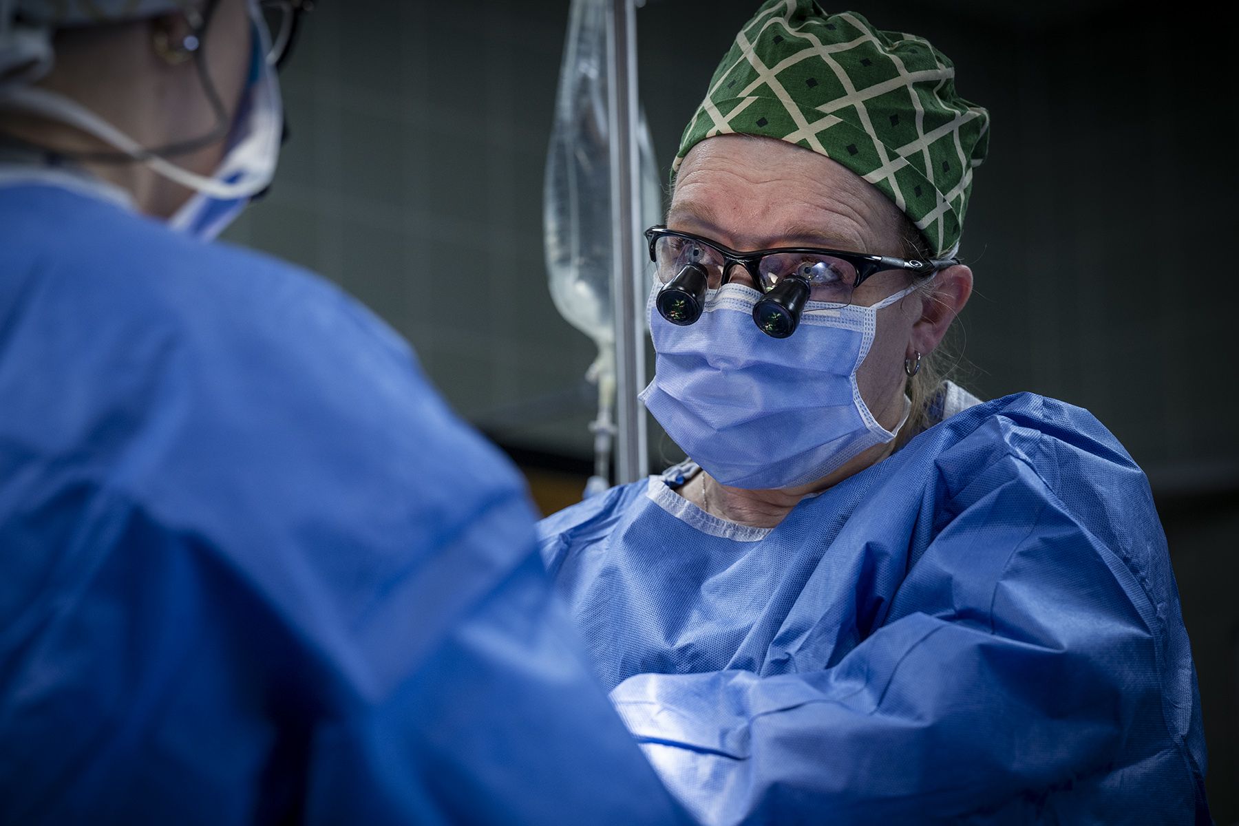 Tammy Richards is looking up from the surgery she’s performing. She’s wearing blue scrubs, gown, mask, a scrub cap and specialized glasses. 