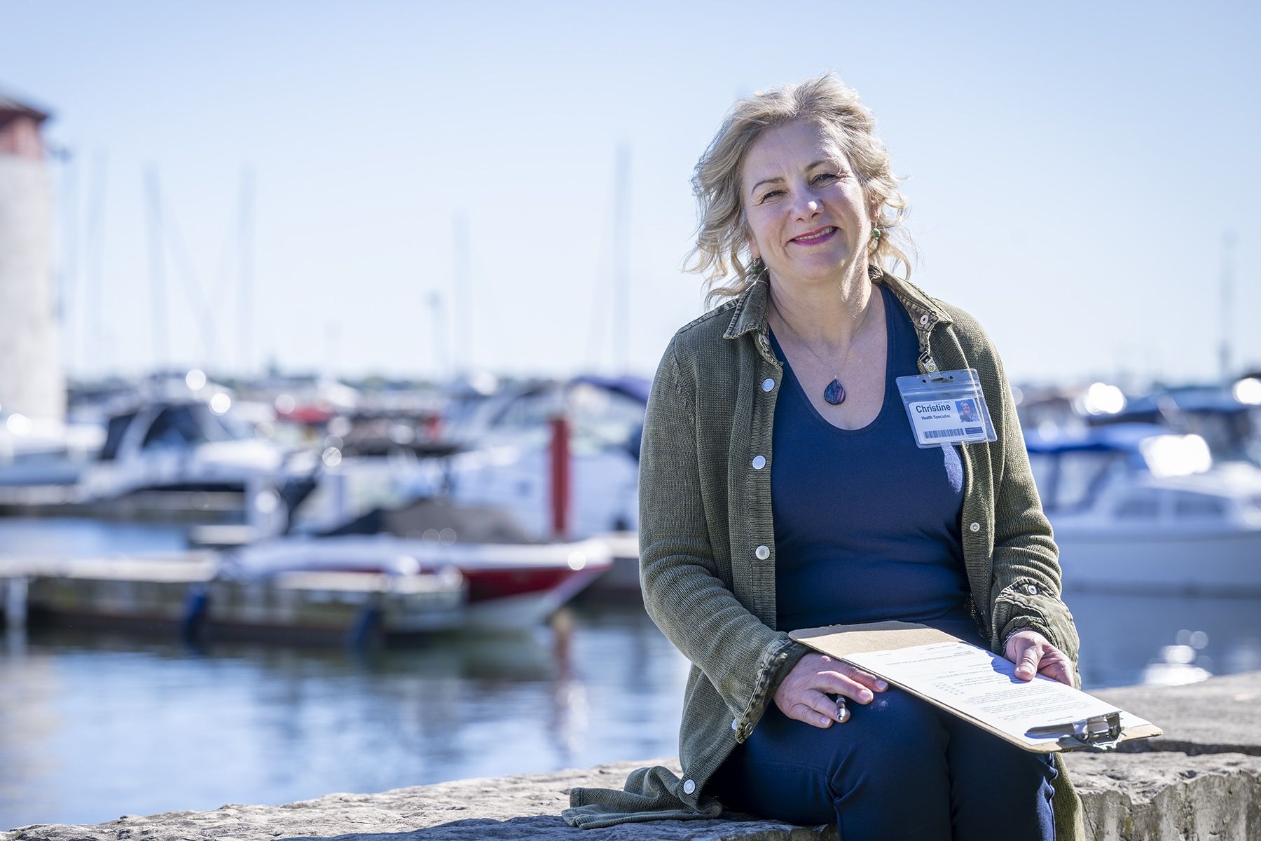 Christine Hennigar is photographed sitting outside of Geaganano Residence. She’s smiling directly into the camera while holding a clipboard. She has light blonde, wavy hair, blue eyes and is wearing a navy blue top with an army green jacket.