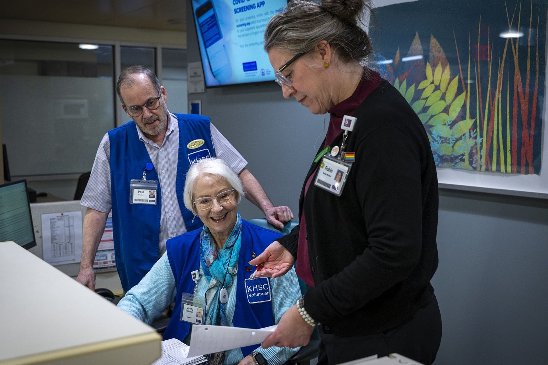 Dorothy Messenger is pictured inside the waiting room at KHSC’s Intensive Care Unit. She has short white hair and wears black framed glasses. She’s wearing a long sleeved, blue sweater with a blue vest with the words KHSC volunteer written on it.