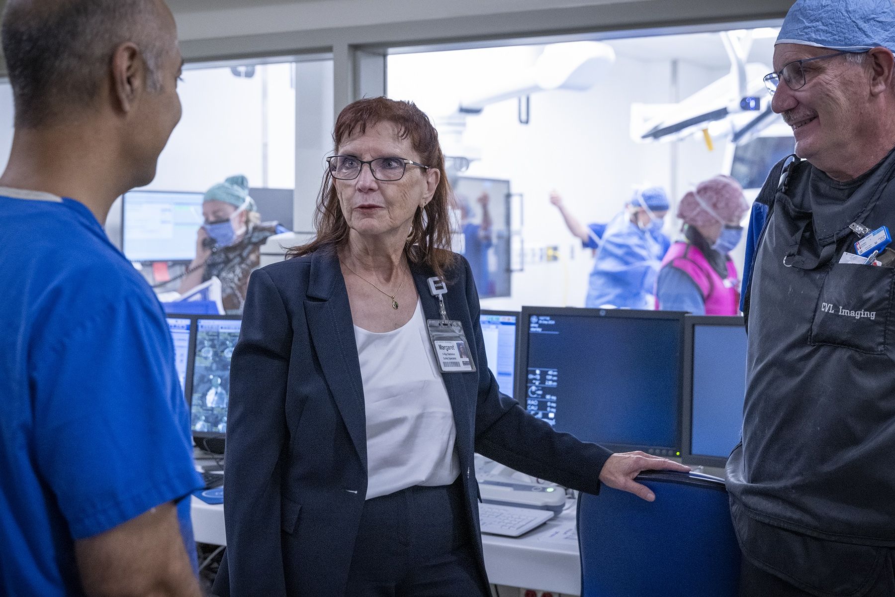 People of KHSC - Margaret Bollen Margaret Bollen is pictured standing in a suite at Kingston General Hospital. She has auburn hair, blue eye hair and wears glasses. She’s wearing a dark navy suit and white top. She’s chatting with physician and a medical radiation technologist.