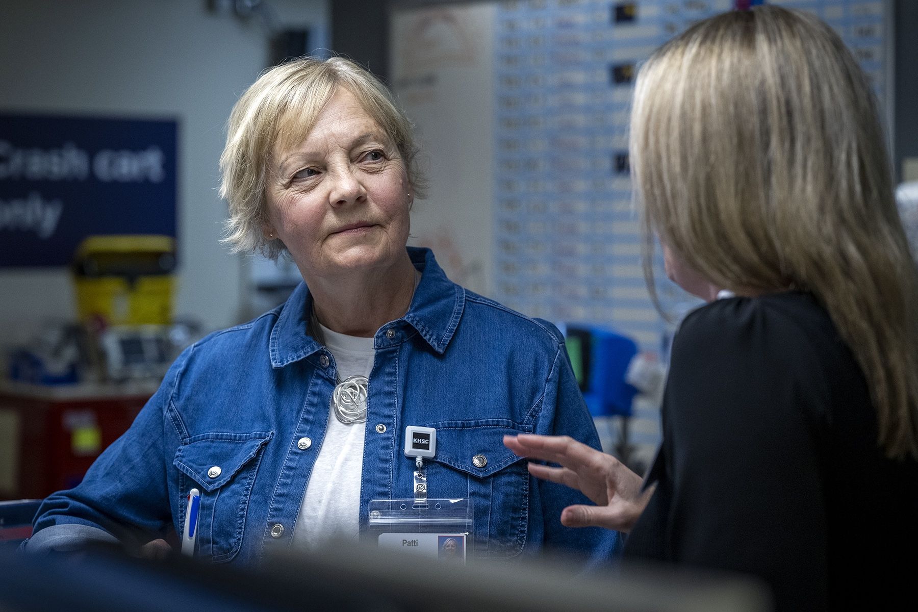 People of KHSC - Patti Cox Patti Cox is pictured leaning against a care desk on a patient care unit, listening as a member of the care team speaks to her. Cox has blue eyes and light blonde hair styled in a pixie cut. She’s wearing a blue denim blouse on top of a white t-shirt. The staff member has their back to the camera, while Cox is facing the camera.