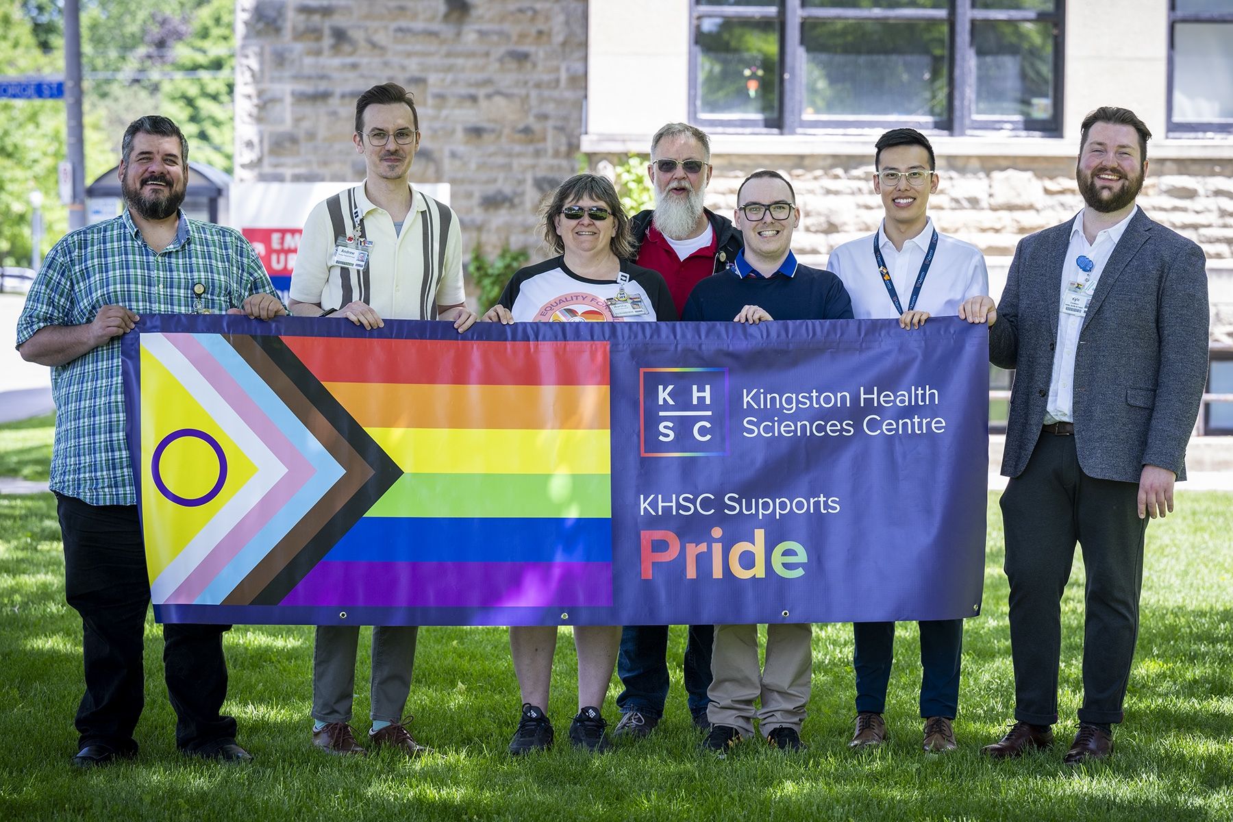 People of KHSC - Pride Staff Community Group Seven members of the Pride Staff Community Group are standing outside Kingston General Hospital, holding a large Progress Pride banner that says Kingston Health Sciences Centre supports Pride.
