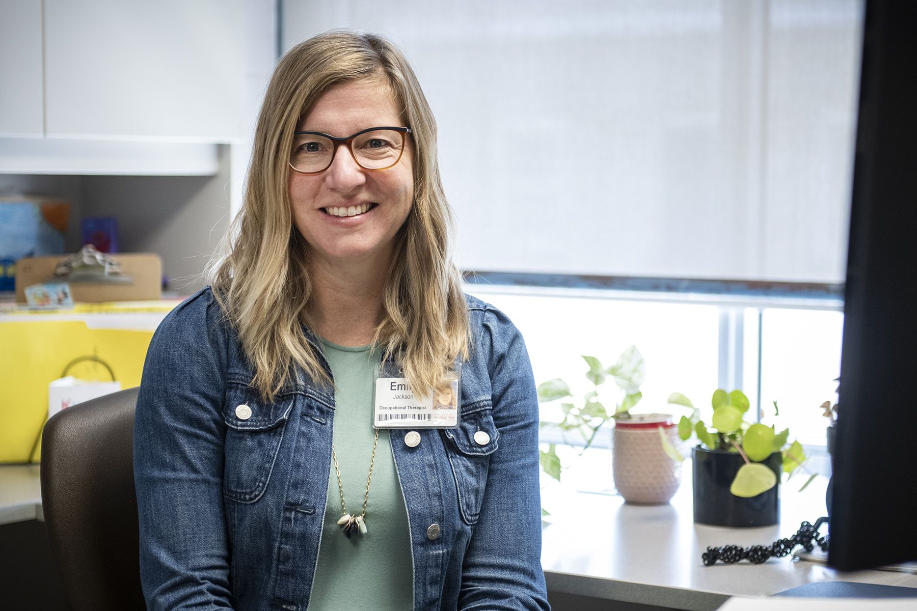 Emily Jackson has shoulder-length blonde hair and blue eyes, and wears glasses. She’s wearing a green top with a jean jacket and is pictured sitting at her desk at KidsInclusive at Hotel Dieu Hospital.