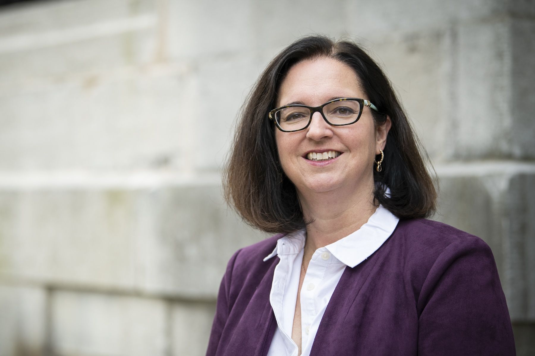 Dr. Jocelyn Garland has shoulder length, dark hair. She's wearing a purple jacket with a white collared shirt. She also wears glasses. She photographed outside the Kingston General Hospital site.