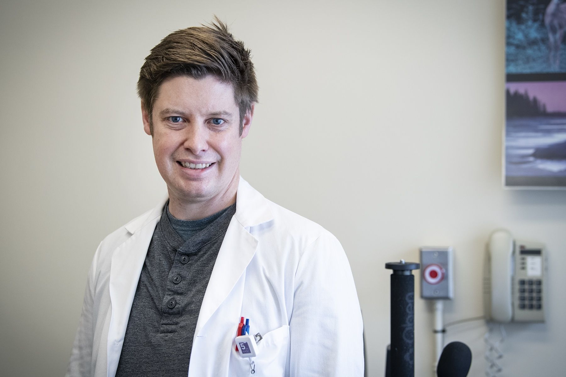 Tim Woodford is pictured inside KHSC’s electroencephalography (EEG) services department. He’s wearing a white lab coat with a grey shirt underneath. He has blue eyes and short, light brown hair.